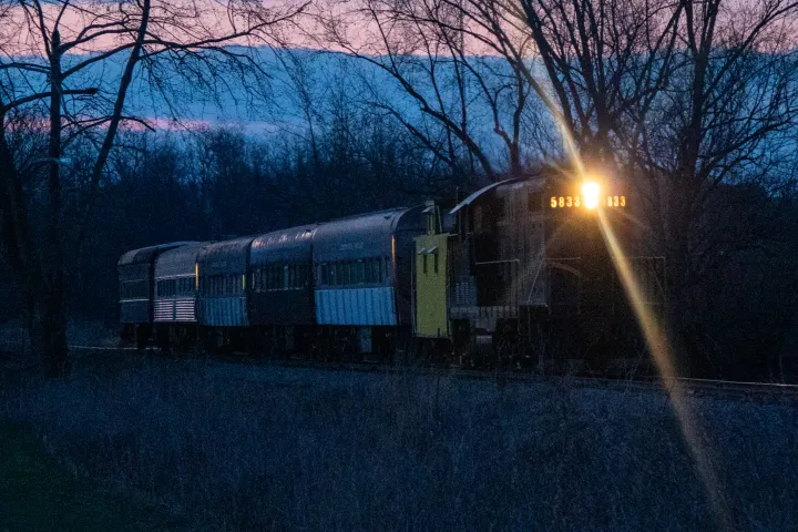 Train with headlight on travels through bare trees at dusk.