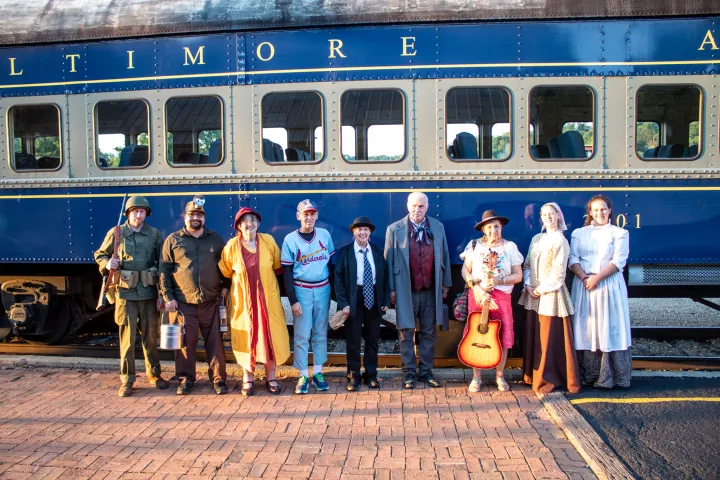People in various vintage costumes stand in front of a blue train car.