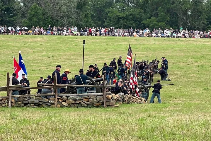 Historical reenactors in Civil War uniforms by a fence with flags, watched by a crowd.