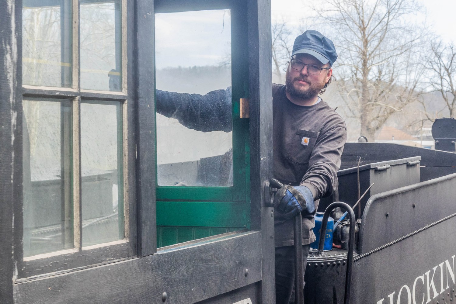 Man in work attire operating a vintage train engine outdoors.