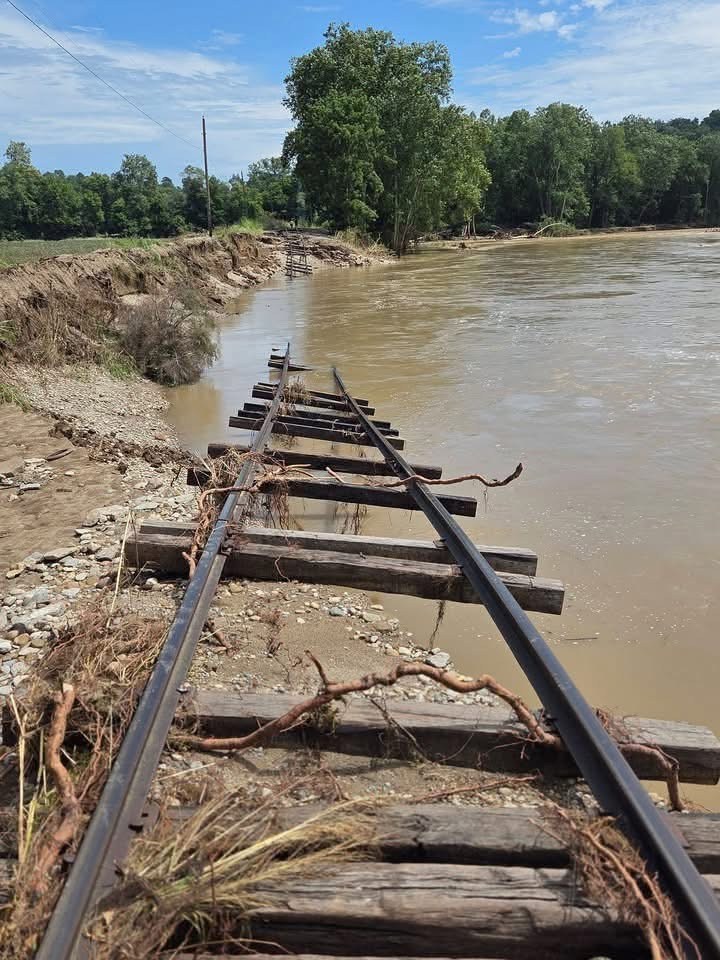 Damaged railway tracks submerged in floodwater near a riverbank.