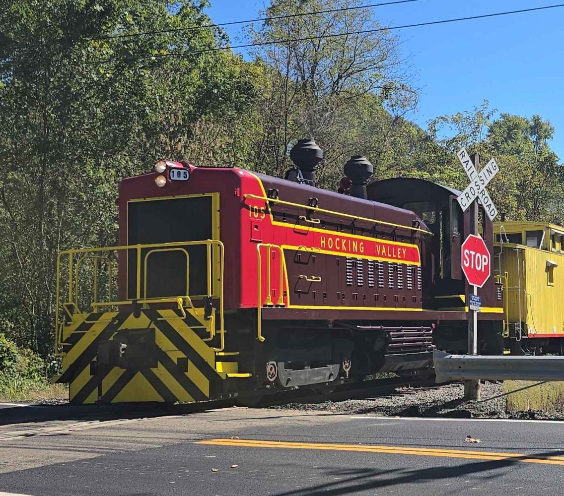 Train crossing a rural road with trees and clear sky in the background.